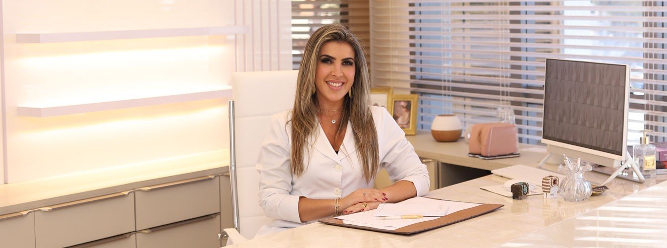 mulher loira sentada na mesa de consultório em frente a computador com os braços sonbre a mesa fundo branco jaleco branco sorrindo simpática